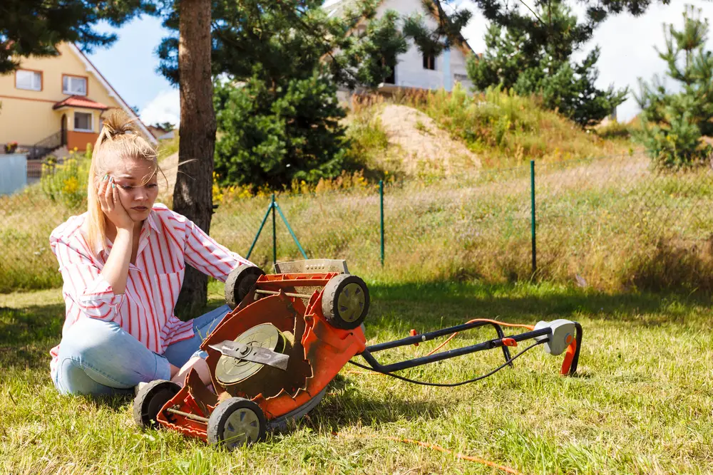 Unhappy woman sitting in the grass with her lawn mower turned over.