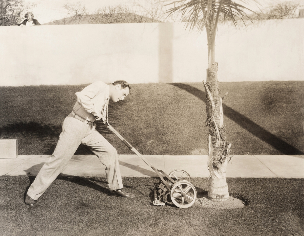 A black-and-white photo of a man using a vintage lawn mower that could use some of today's technologies.