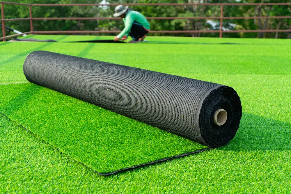 A person laying down artificial grass in the background with a roll in the foreground.