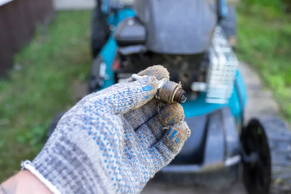 A person holding a spark plug in front of a blurry lawn mower.