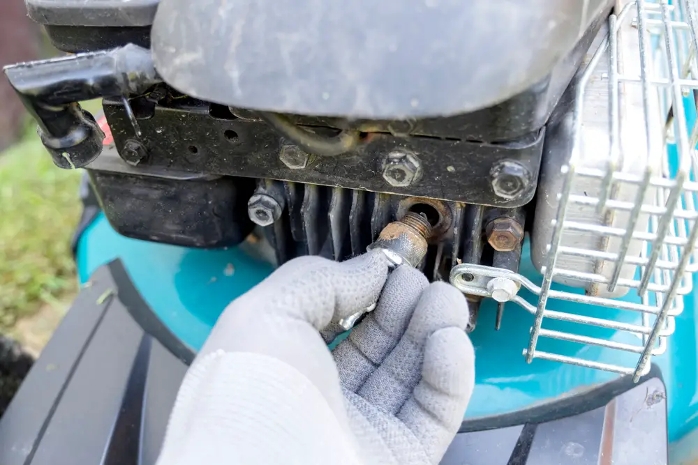 A person pulling a spark plug out of a turquoise lawn mower.