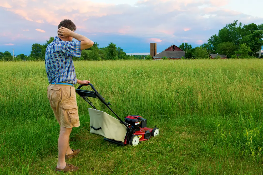 A man scratching the back of his head while looking at all the tall grass he has to cut with his lawn mower.