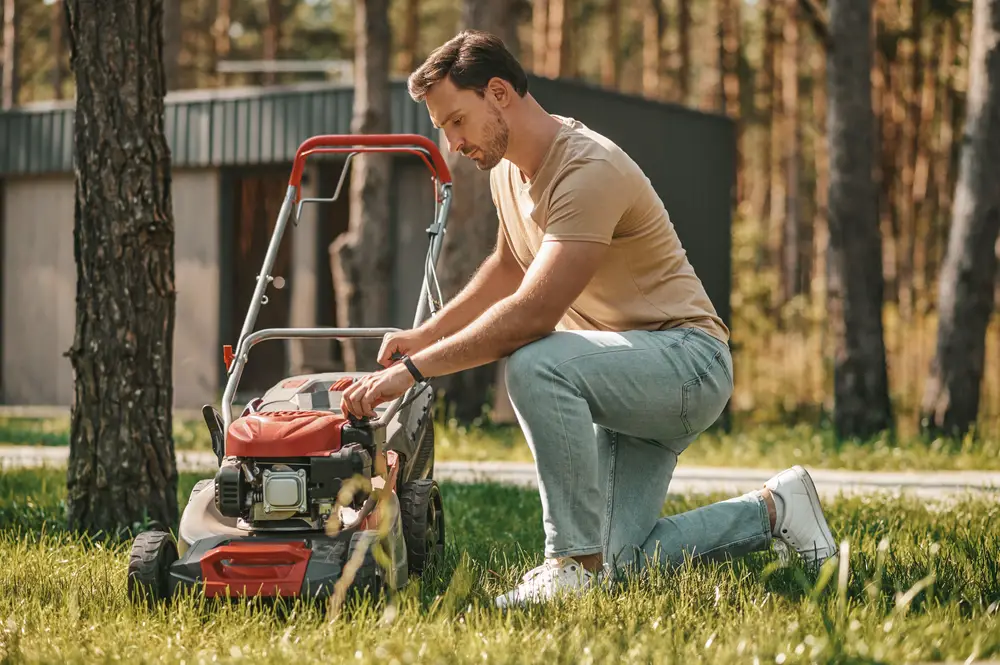 A man in jeans kneeling by a lawn mower while outside.
